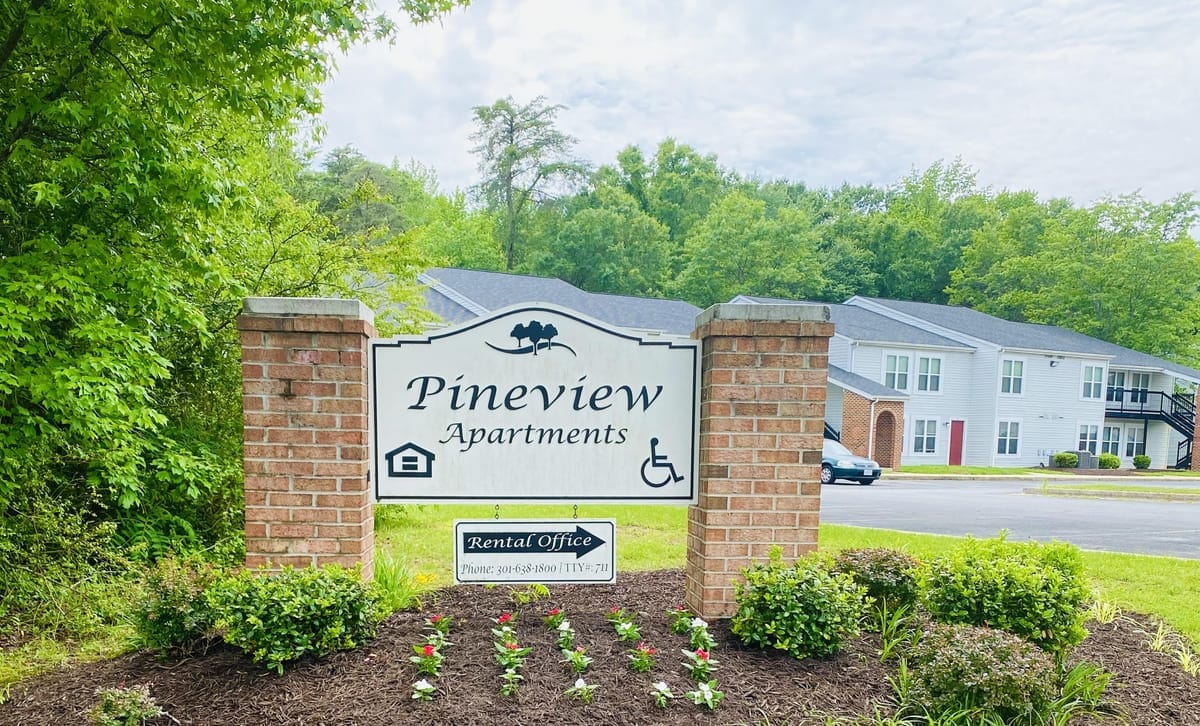 The Pineview Apartments entrance sign on brick pillars, with a planted flower bed in front and a two-story apartment building and trees behind